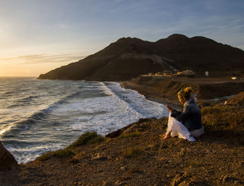 Man sitting on beach by sea against sky during sunset