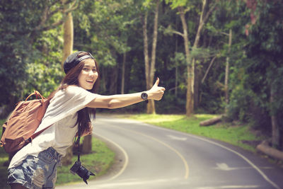 Portrait of smiling woman hitchhiking on road against trees