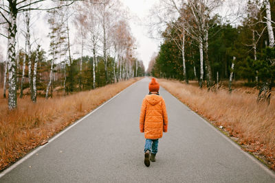 Girl in orange coat walks along the road in fall forest. thinking and mental health. autumn vibes