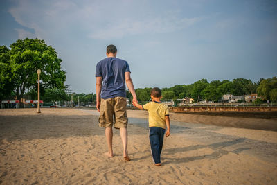 Rear view full length of father with son walking at sandy beach against sky