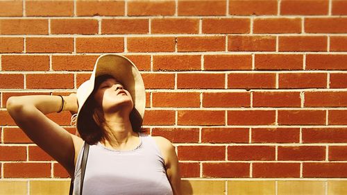 Woman wearing hat standing with head back against brick wall on sunny day