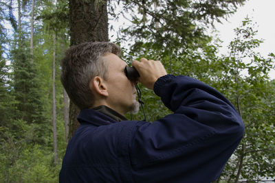 Side view of man looking through binoculars while standing in forest