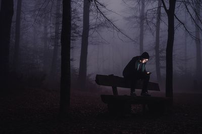 Man using mobile phone while sitting on bench in forest at night