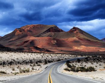 Country road leading towards mountains against cloudy sky