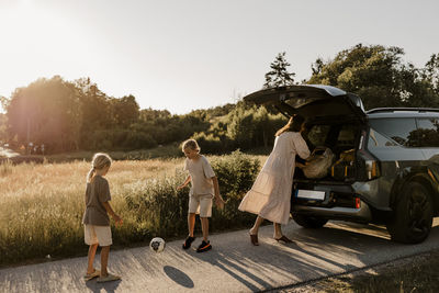 Mother near car trunk with boys playing with soccer ball on driveway by meadow at sunny day