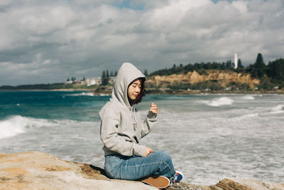 Full length of woman sitting on rock at beach against sky