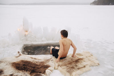Rear view of shirtless boy sitting with legs in cold water at frozen lake