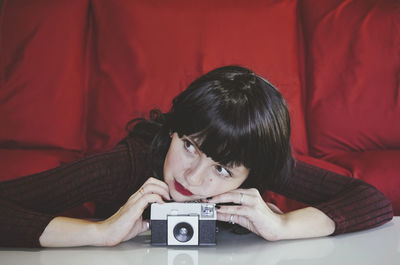 Thoughtful woman leaning on analog camera in front of red couch