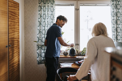 Male caregiver looking at senior woman while watering plants at home