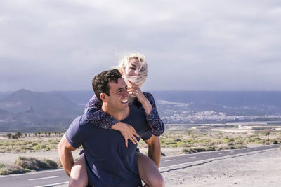 Man piggybacking woman by road against sky