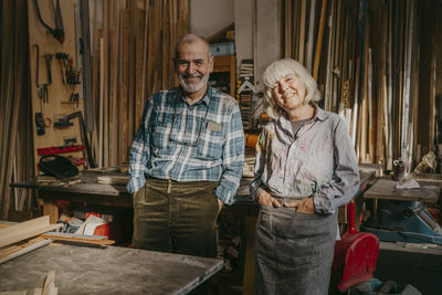 Portrait of happy male and female carpenters with hands in pockets standing at workshop