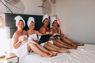 Four girls friends in white towels sitting on the bed having fun in hotel. vacation bloggers 