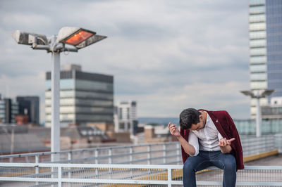 Man photographing woman using mobile phone in city against sky