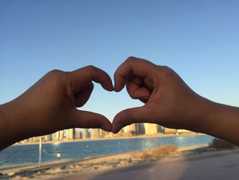 Cropped hands making heart shape with river in background against clear sky