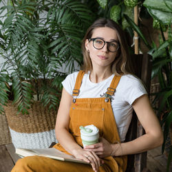 Portrait of woman holding coffee while sitting on chair against plants