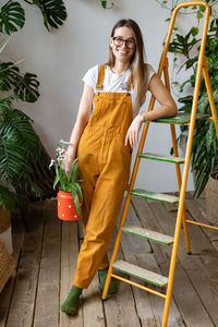 Portrait of smiling woman with potted plant standing by ladder at home