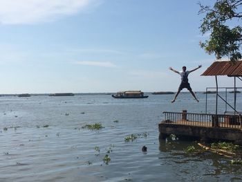 Full length of man jumping with arms outstretched on jetty over sea against sky