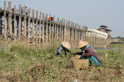 Woman with wicker basket while crouching on field against bridge