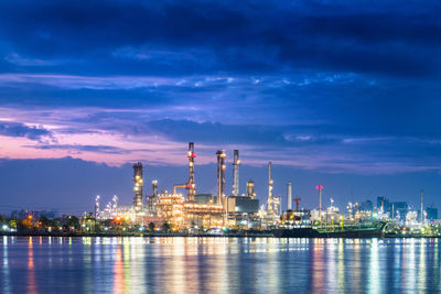 Illuminated factory by sea against sky at night