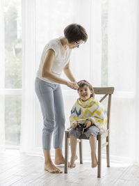 Mother cuts her son's hair by herself. boy sits, covered with cloth, and holds pair of scissors. 