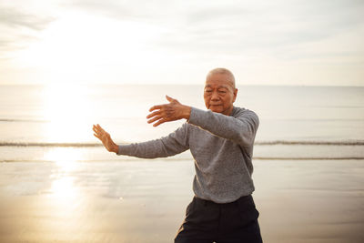 Senior man exercising while standing at beach against sky during sunset