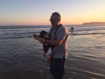 Grandfather holding child in baby carriage while standing at beach during sunset