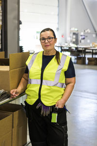 Portrait of confident mature female worker standing by cart in distribution warehouse