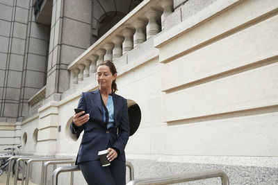 Businesswoman with coffee cup using mobile phone while leaning on railing of bicycle parking lot