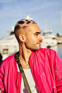 Eccentric young male with unusual haircut sitting on marina with yachts and looking away with positive glance