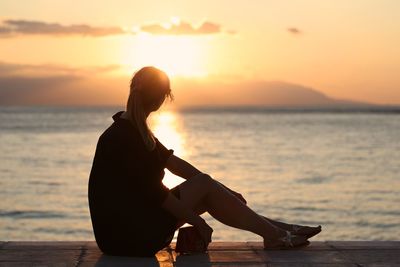 Silhouette woman sitting on beach against sky during sunset