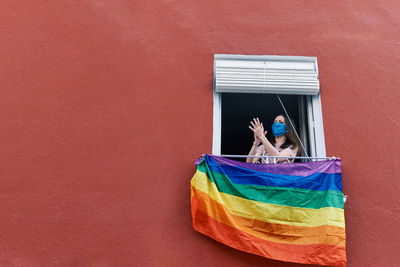 Young woman in a mask clapping from the window with the gay flag on a red background