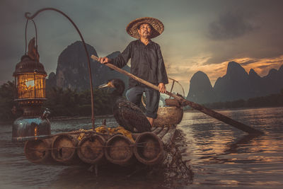 Portrait of traditional fisherman by cormorants on wooden raft over river at sunset