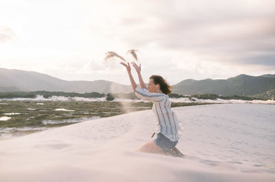 Side view of woman throwing sand while kneeling at beach