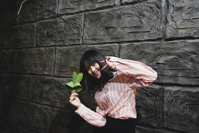 Portrait of young woman holding leaves while gesturing peace sign by wall at night