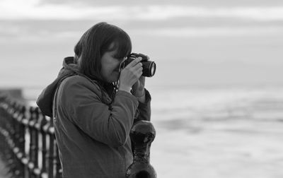 Side view of woman photographing sea against cloudy sky