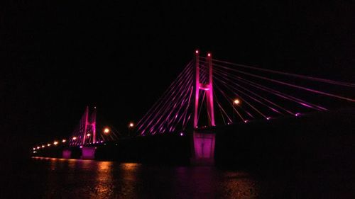 Illuminated suspension bridge over river against sky at night