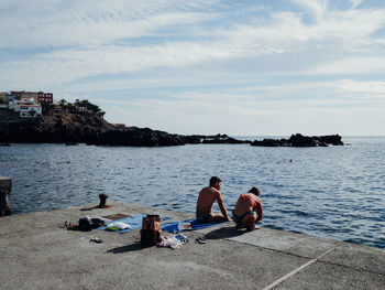 People sitting on shore by sea against sky