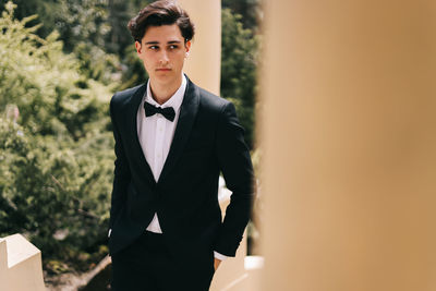 A beautiful young man, the groom in an elegant wedding suit, stands posing in the city's old park