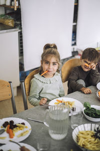 Portrait of playful girl holding carrot slice in mouth while sitting at dining table during lunch time