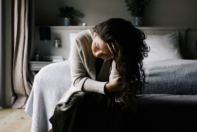 Emotionally depressed woman sitting with hand in hair against bed at home