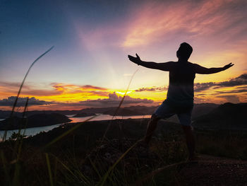 Rear view of man standing with arms outstretched on land against sky during sunset