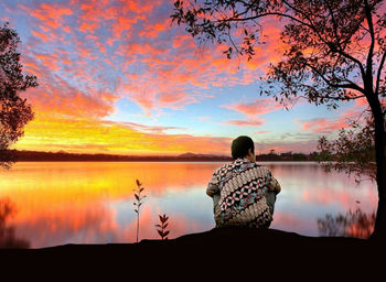 Rear view of man sitting by lake against sky during sunset