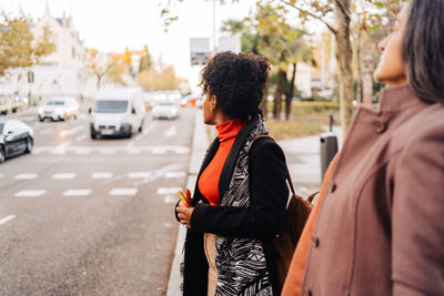 Side view of selective focus of ethnic female and hispanic lady in outerwear looking away while standing near asphalt road in city