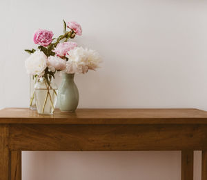 Pink and white peonies in glass and ceramic vases on oak wooden table against neutral wall 