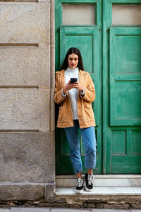 Full length of man using mobile phone while standing against wall