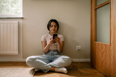 Girl using smart phone while sitting cross-legged on floor against wall at home