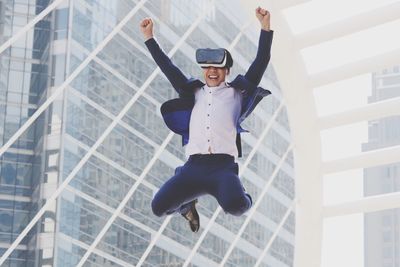 Low angle view of smiling businessman enjoying while using vr glasses against building