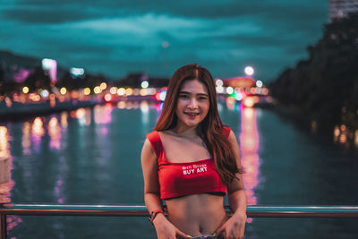 Portrait of smiling young woman standing against illuminated water
