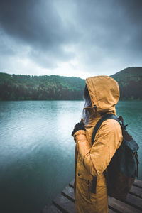 Young woman wearing raincoat while standing on pier by lake during rainy season