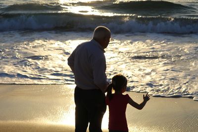 Rear view of girl with grandfather standing on shore at beach